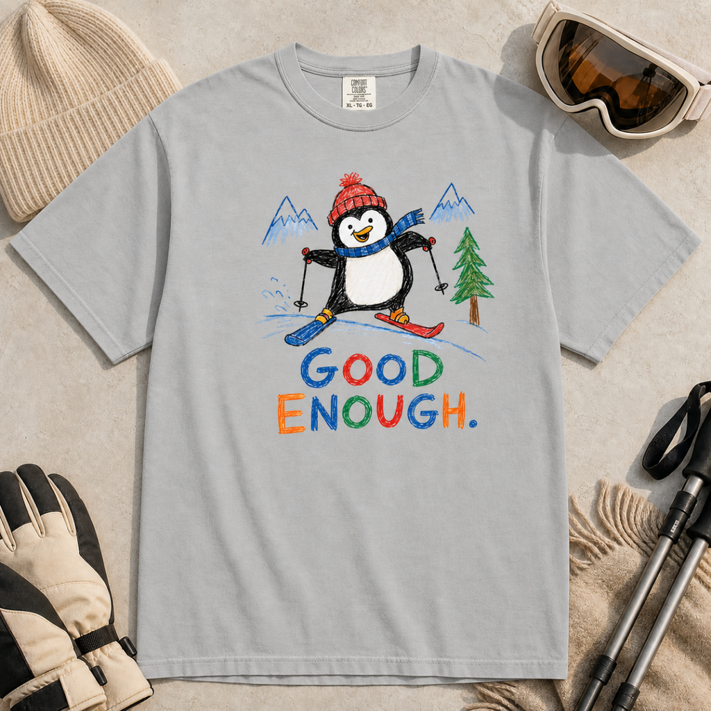 Flat lay of a granite gray t-shirt featuring a childlike crayon-style ski design with a happy penguin skiing downhill, mountains, a pine tree, and colorful “Good Enough.” text.
