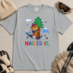 Flat lay of a granite gray t-shirt featuring a playful hand-drawn bear ski crash graphic with mountains, snow, a pine tree, and “Nailed It.” text.