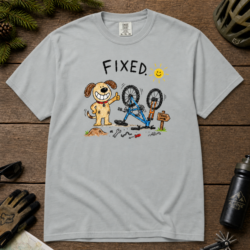 Flat lay of a granite gray t-shirt featuring a childlike crayon-style bike repair design with a smiling dog, upside-down mountain bike, tools, and “FIXED.” text.