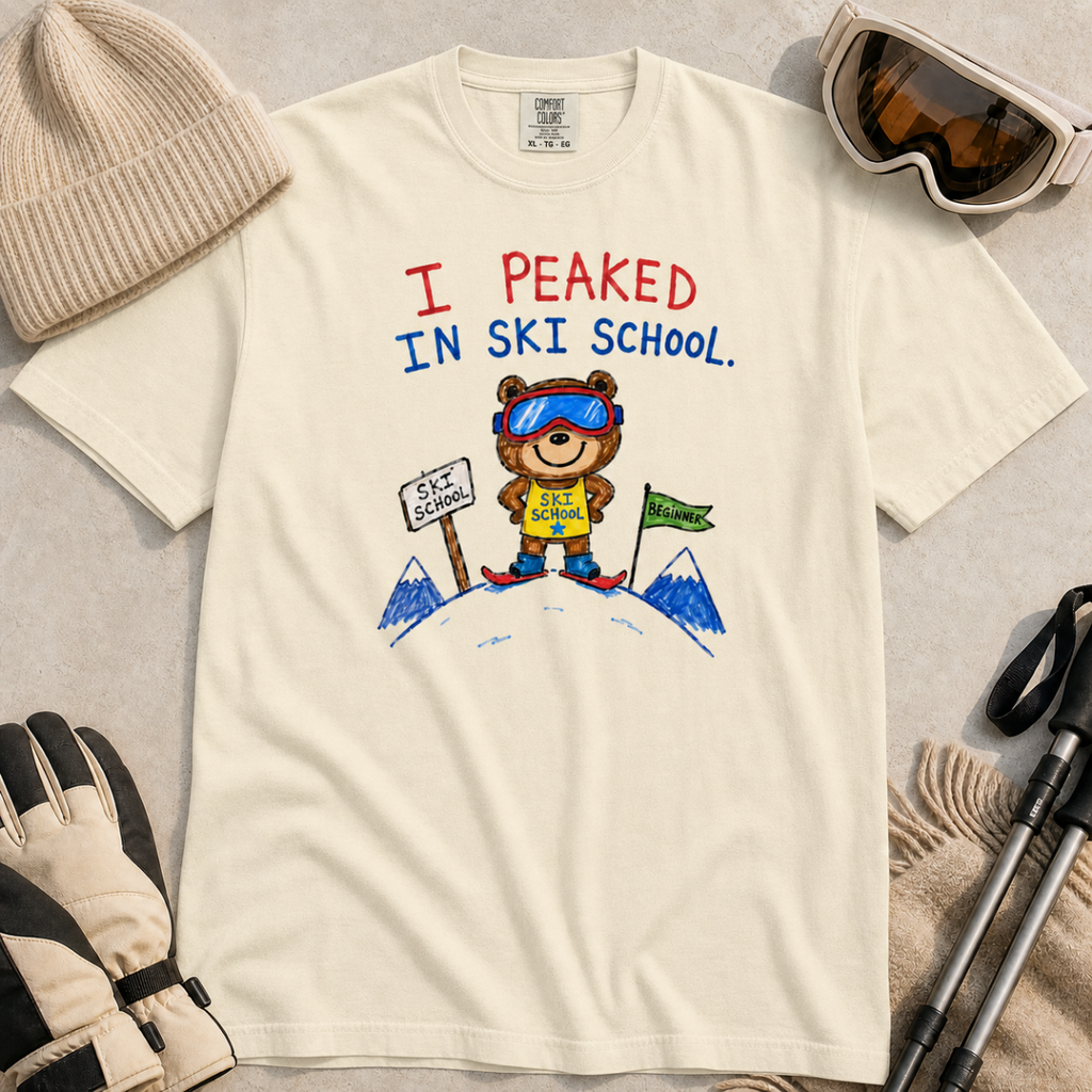 Flat lay of an ivory white t-shirt featuring a childlike crayon-style ski design with a proud bear in ski school gear and “I Peaked in Ski School.” text.