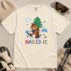 Flat lay of an ivory white t-shirt featuring a playful hand-drawn bear ski crash graphic with mountains, snow, a pine tree, and “Nailed It.” text.