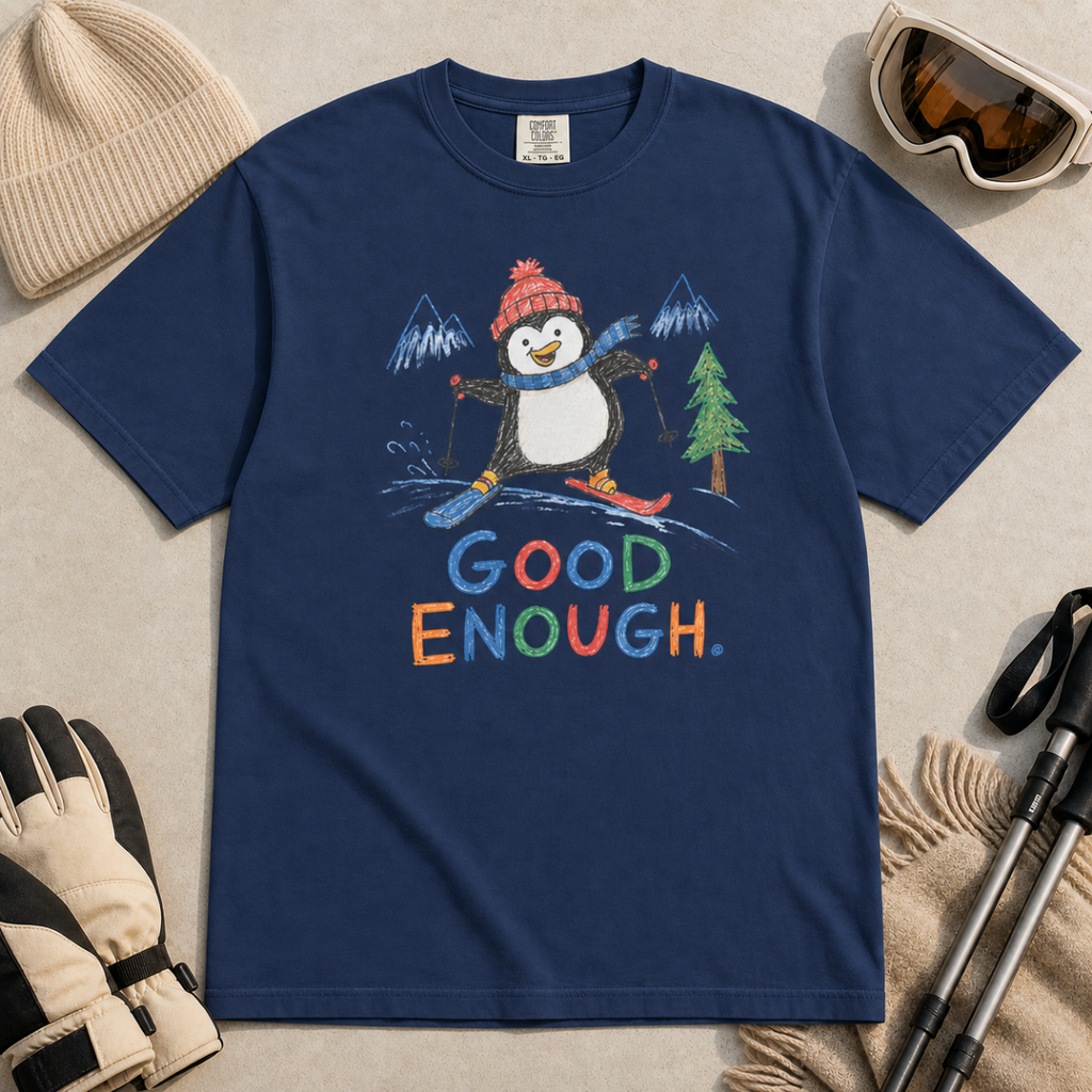 Flat lay of a navy t-shirt featuring a childlike crayon-style ski design with a happy penguin skiing downhill, mountains, a pine tree, and colorful “Good Enough.” text.