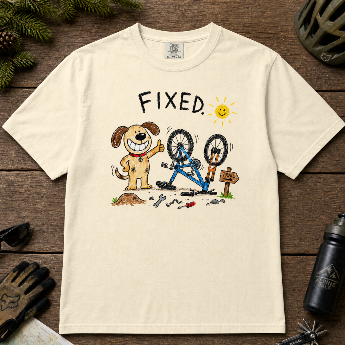 Flat lay of an ivory white t-shirt featuring a childlike crayon-style bike repair design with a smiling dog, upside-down mountain bike, tools, and “FIXED.” text.