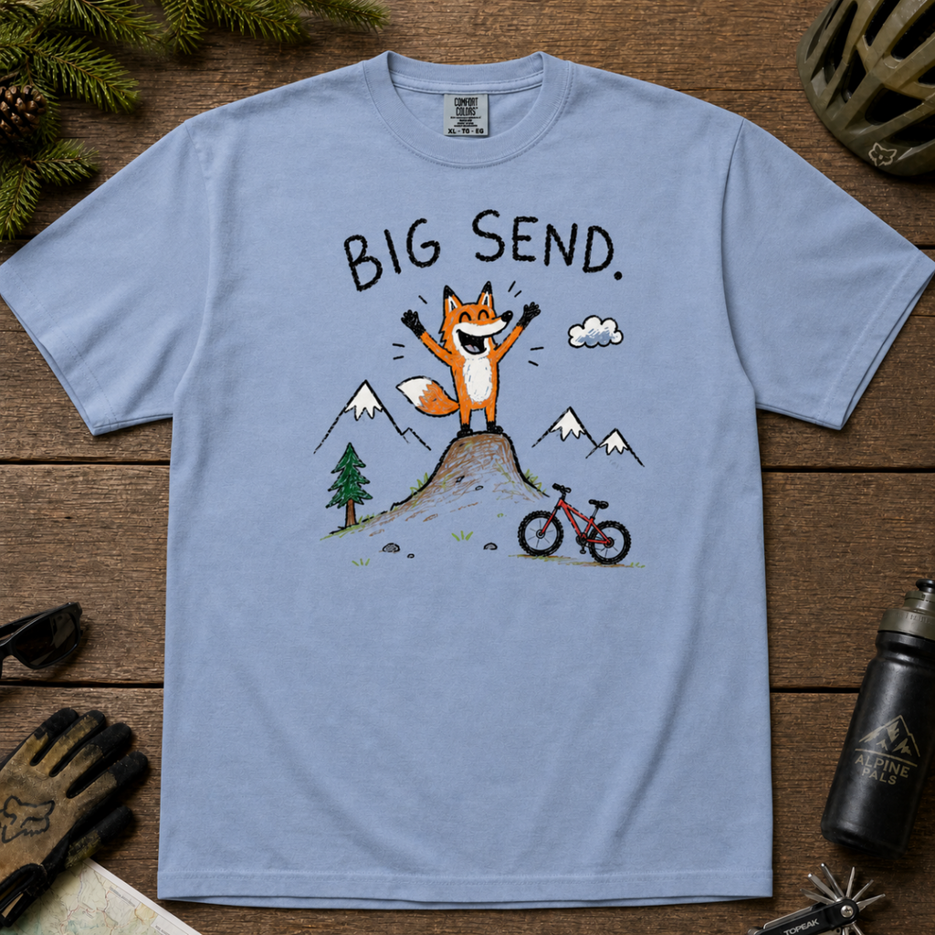 Flat lay of a washed denim blue t-shirt featuring a playful hand-drawn fox mountain bike graphic with mountains, a small bike, and “Big Send.” text.