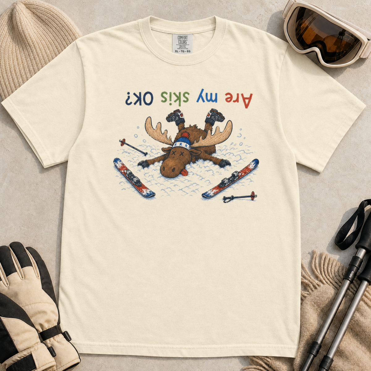 Flat lay of an ivory white t-shirt featuring a humorous upside-down “Are My Skis OK?” design with a moose face-planted in the snow among scattered skis and poles.