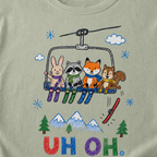 Close-up of a bay green t-shirt with a childlike crayon-style ski design showing four animals on a chairlift, a loose red ski falling below, and colorful “UH OH.” text.