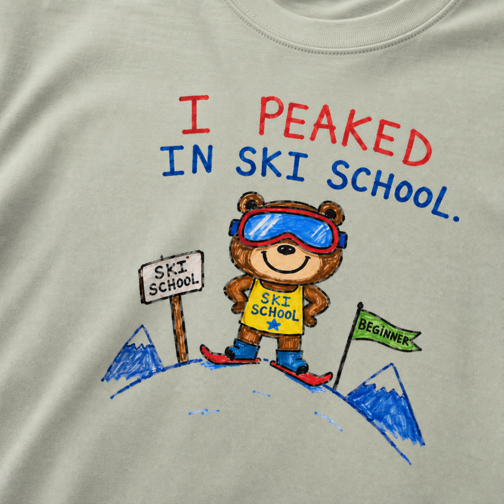 Close-up of a bay green t-shirt with a playful hand-drawn bear at ski school, beginner flag, mountain details, and “I Peaked in Ski School.” text.