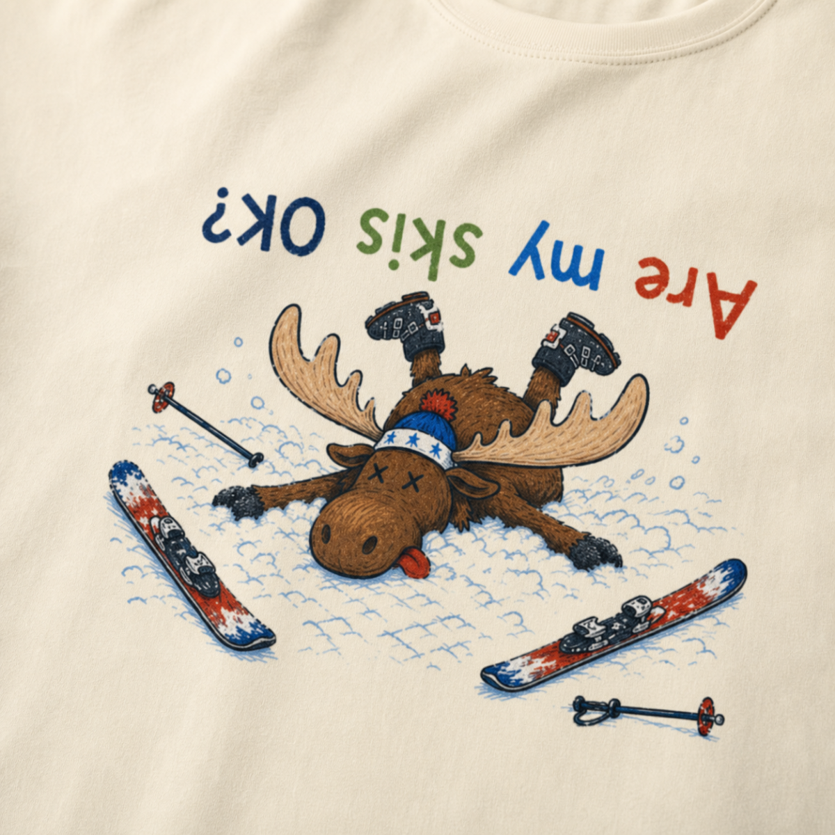 Close-up of an ivory white t-shirt showing a crashed moose lying upside down in the snow with loose ski gear and colorful “Are My Skis OK?” text.