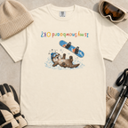 Flat lay of an ivory white t-shirt featuring an upside-down “Is My Snowboard OK?” design with a dazed otter lying in the snow beneath a loose snowboard.