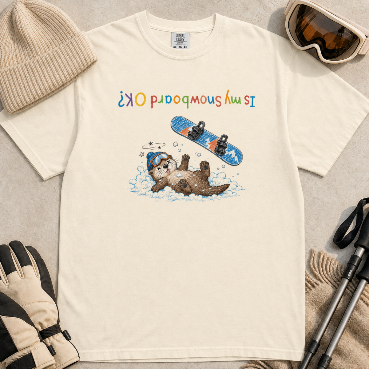 Flat lay of an ivory white t-shirt featuring an upside-down “Is My Snowboard OK?” design with a dazed otter lying in the snow beneath a loose snowboard.