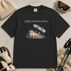 Flat lay of a black t-shirt featuring an upside-down “Is My Snowboard OK?” design with a dazed otter lying in the snow beneath a loose snowboard.