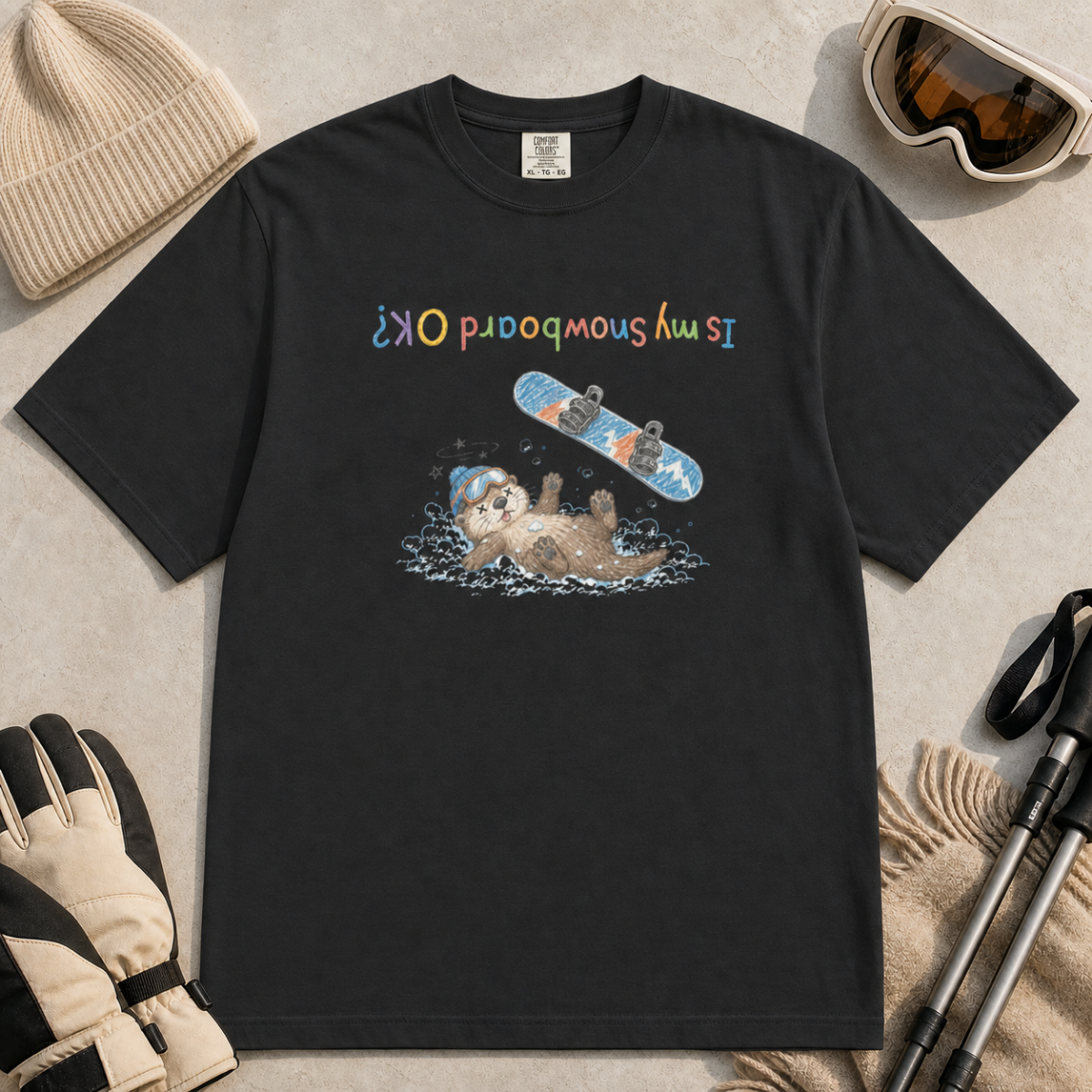 Flat lay of a black t-shirt featuring an upside-down “Is My Snowboard OK?” design with a dazed otter lying in the snow beneath a loose snowboard.