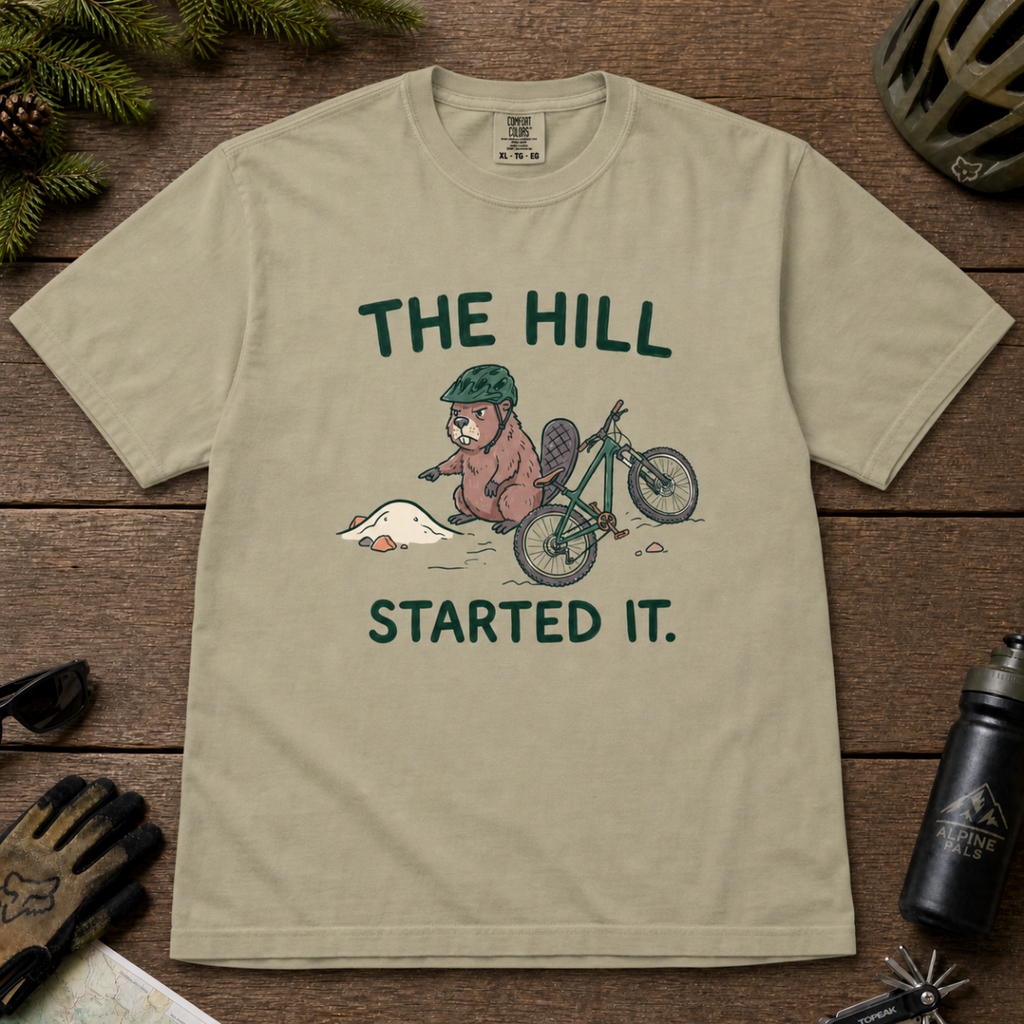 Flat lay of a khaki t-shirt with a humorous mountain biking design showing an annoyed beaver blaming a dirt hill beside its bike.