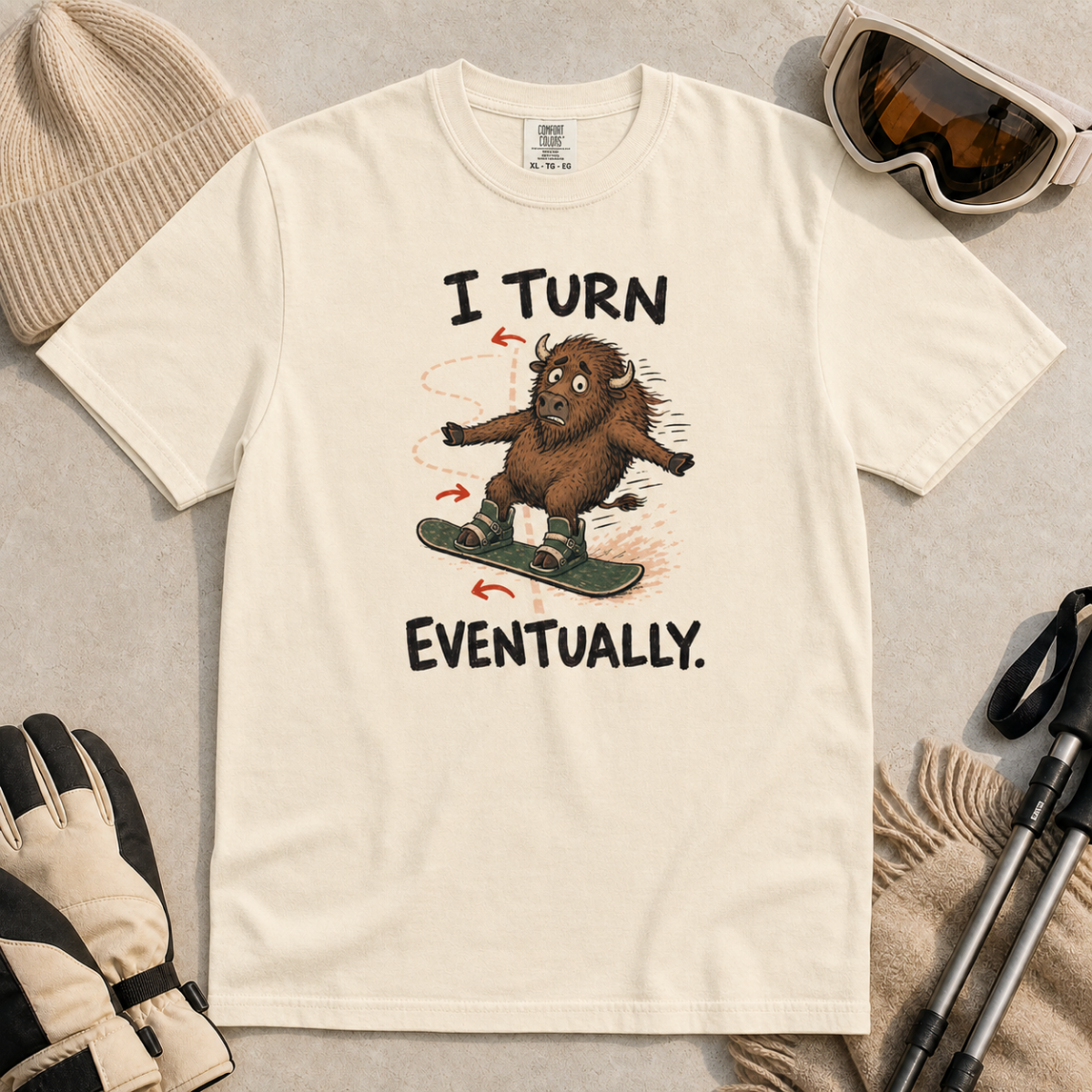 Flat lay of an ivory white t-shirt featuring a nervous bison snowboarding along a winding path with “I Turn Eventually.” text.