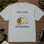 Flat lay of a granite gray t-shirt with a playful mountain hiking design showing a small backpacking hamster approaching a tall peak.