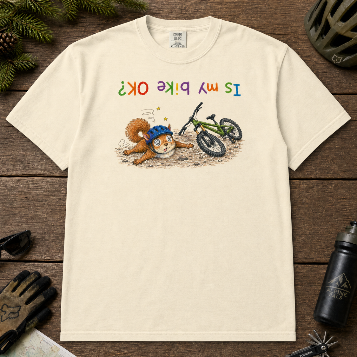 Flat lay of an ivory white t-shirt featuring a humorous squirrel mountain bike crash design with an upside-down “Is My Bike OK?” message.