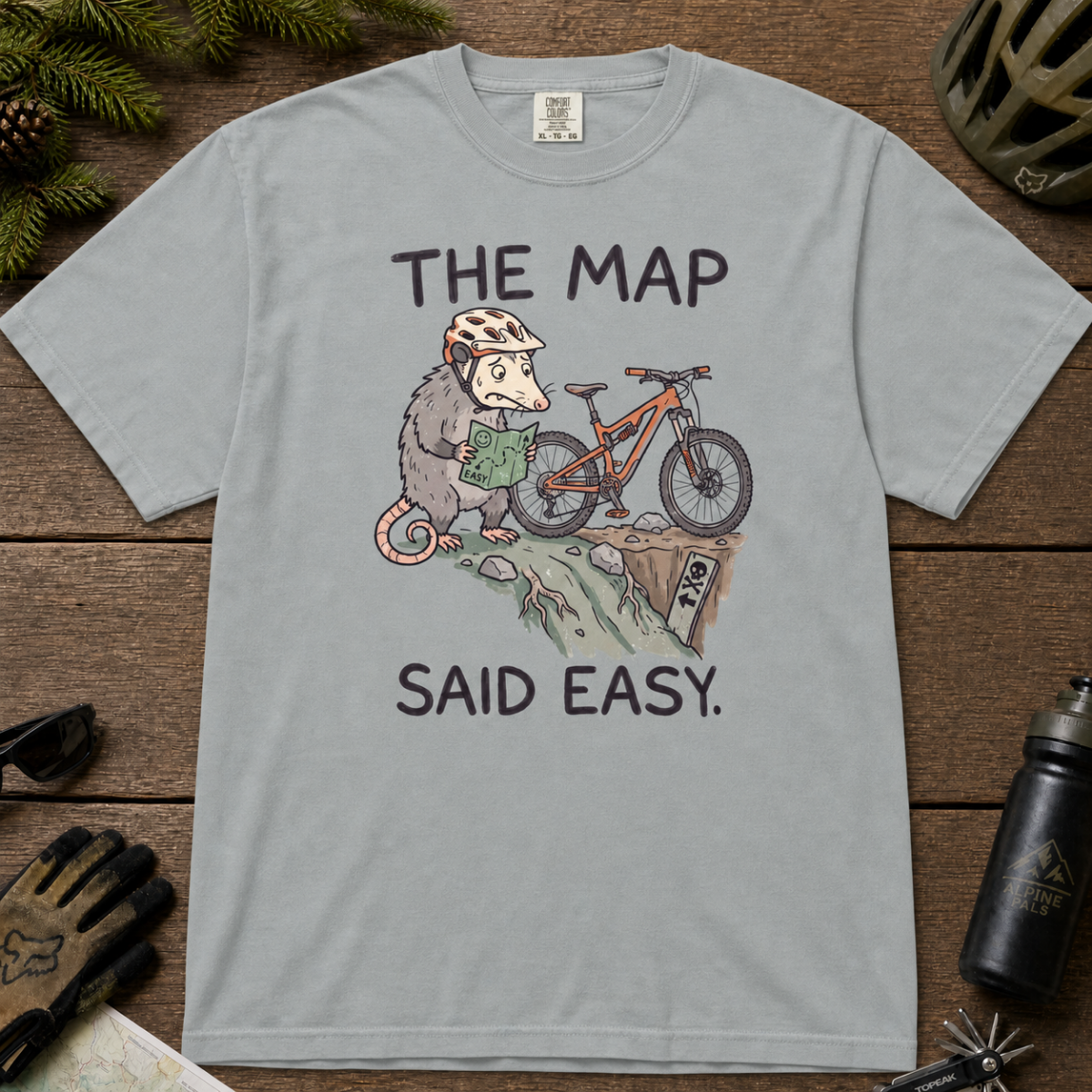 Flat lay of a granite gray t-shirt with a humorous mountain biking design showing a nervous opossum, an orange bike, and a dangerously steep trail.