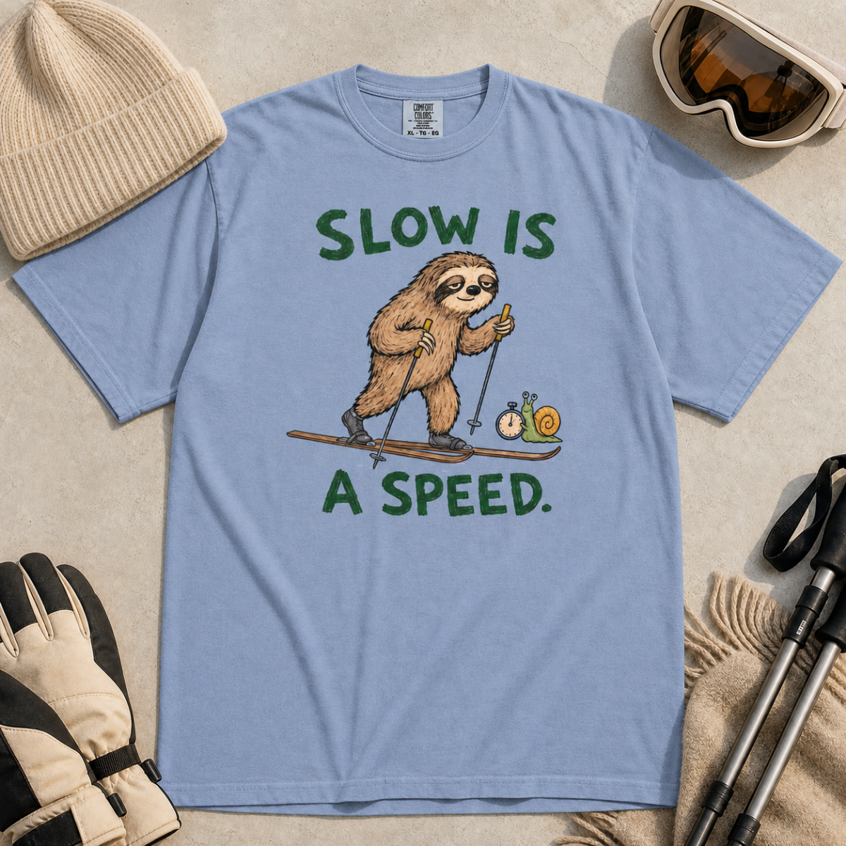 Flat lay of a washed denim blue t-shirt with a humorous sloth skiing graphic, a stopwatch-holding snail, and bold green “Slow Is a Speed.” text.