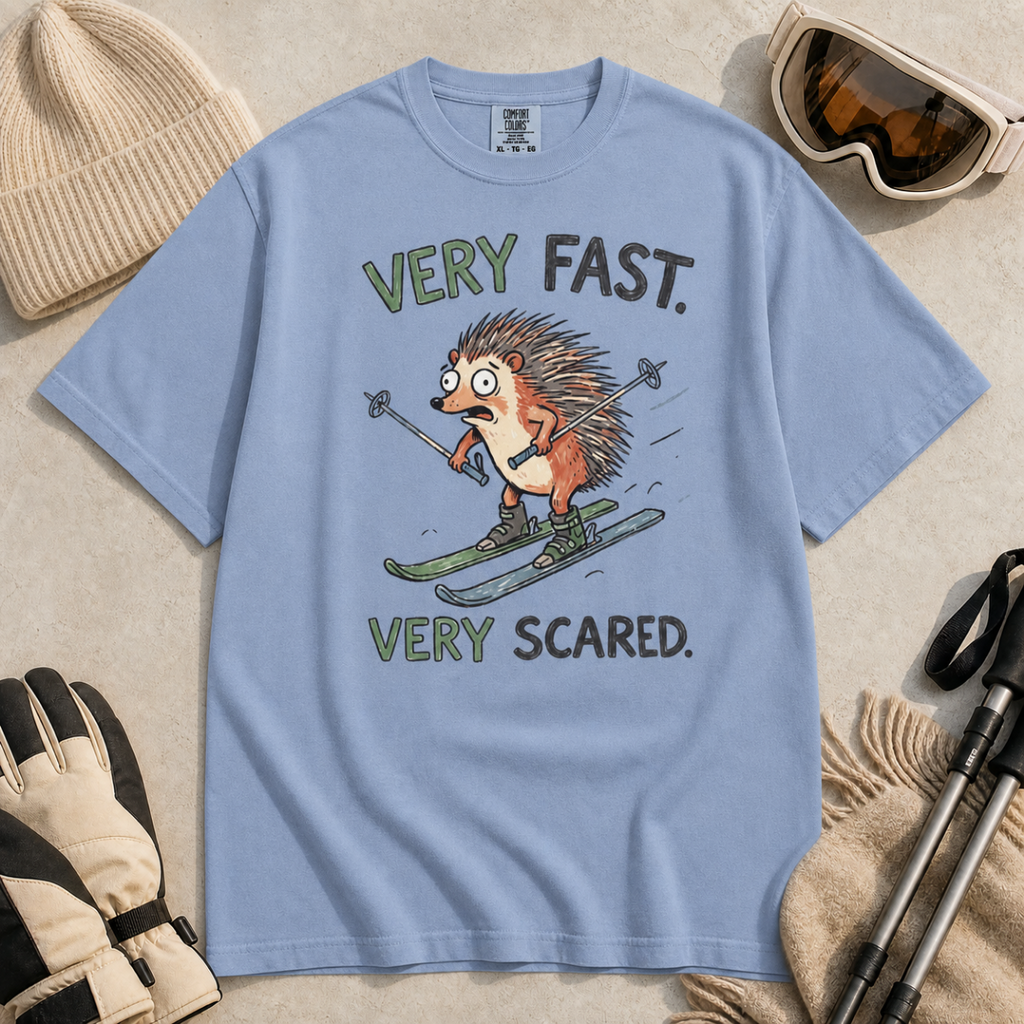 Flat lay of a washed denim blue t-shirt featuring a frightened hedgehog skiing at high speed with “Very Fast. Very Scared.” text, styled with winter accessories.