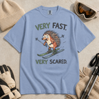 Flat lay of a washed denim blue t-shirt featuring a frightened hedgehog skiing at high speed with “Very Fast. Very Scared.” text, styled with winter accessories.