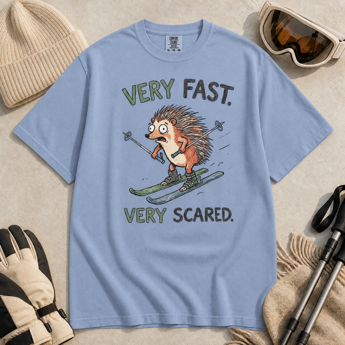 Flat lay of a washed denim blue t-shirt featuring a frightened hedgehog skiing at high speed with “Very Fast. Very Scared.” text, styled with winter accessories.