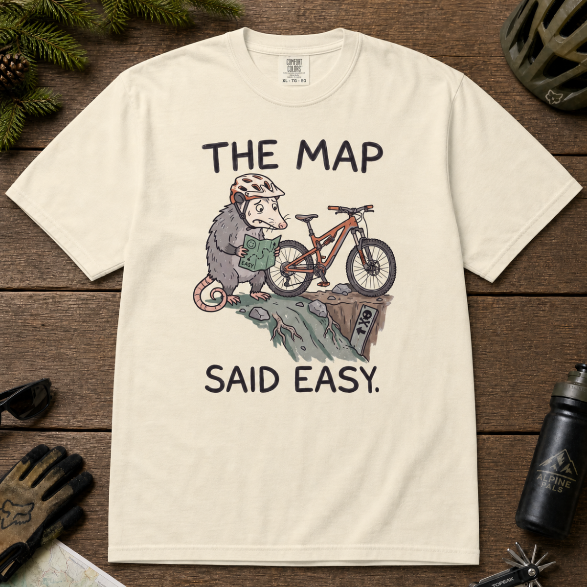 Flat lay of an ivory white t-shirt with a humorous mountain biking design showing a nervous opossum, an orange bike, and a dangerously steep trail.