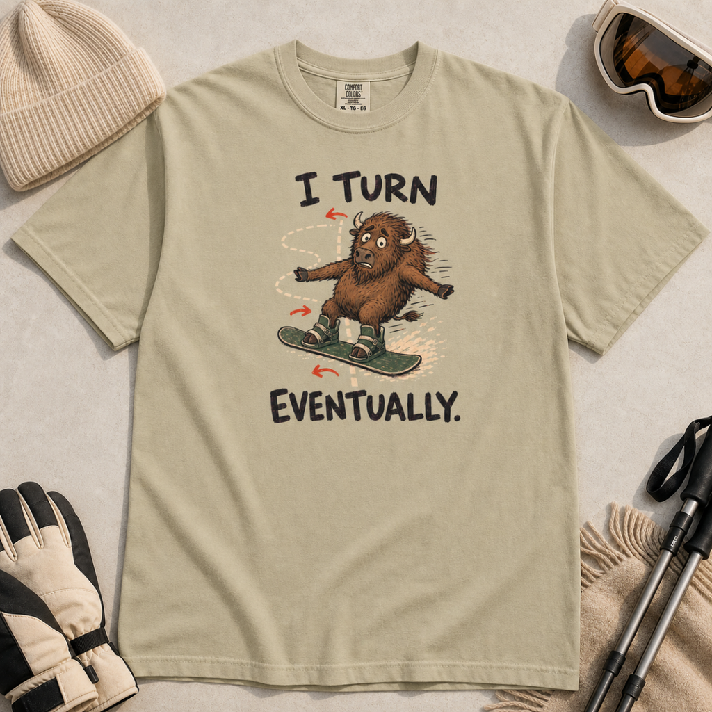 Flat lay of a khaki t-shirt featuring a nervous bison snowboarding along a winding path with “I Turn Eventually.” text.