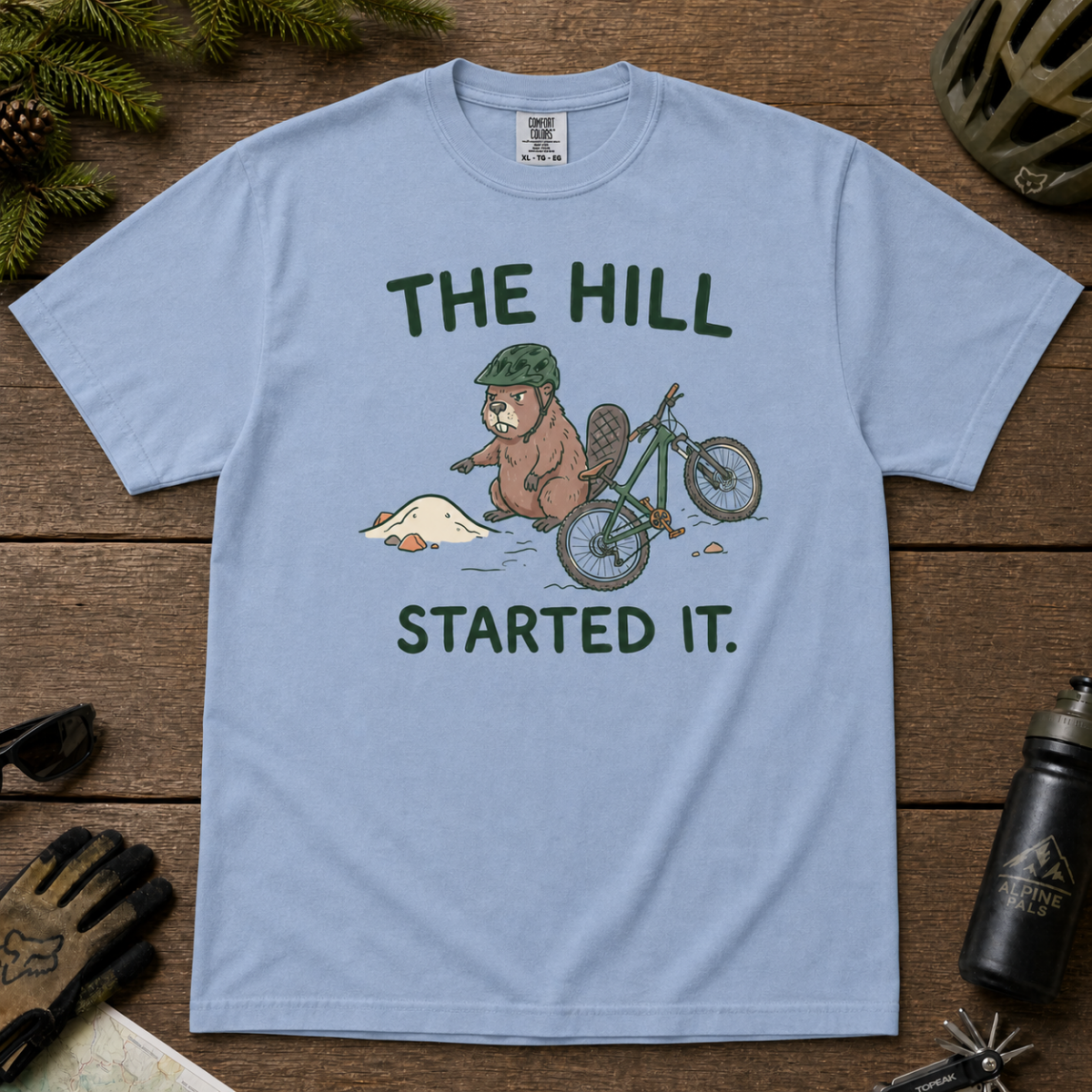 Flat lay of a washed denim blue t-shirt with a humorous mountain biking design showing an annoyed beaver blaming a dirt hill beside its bike.