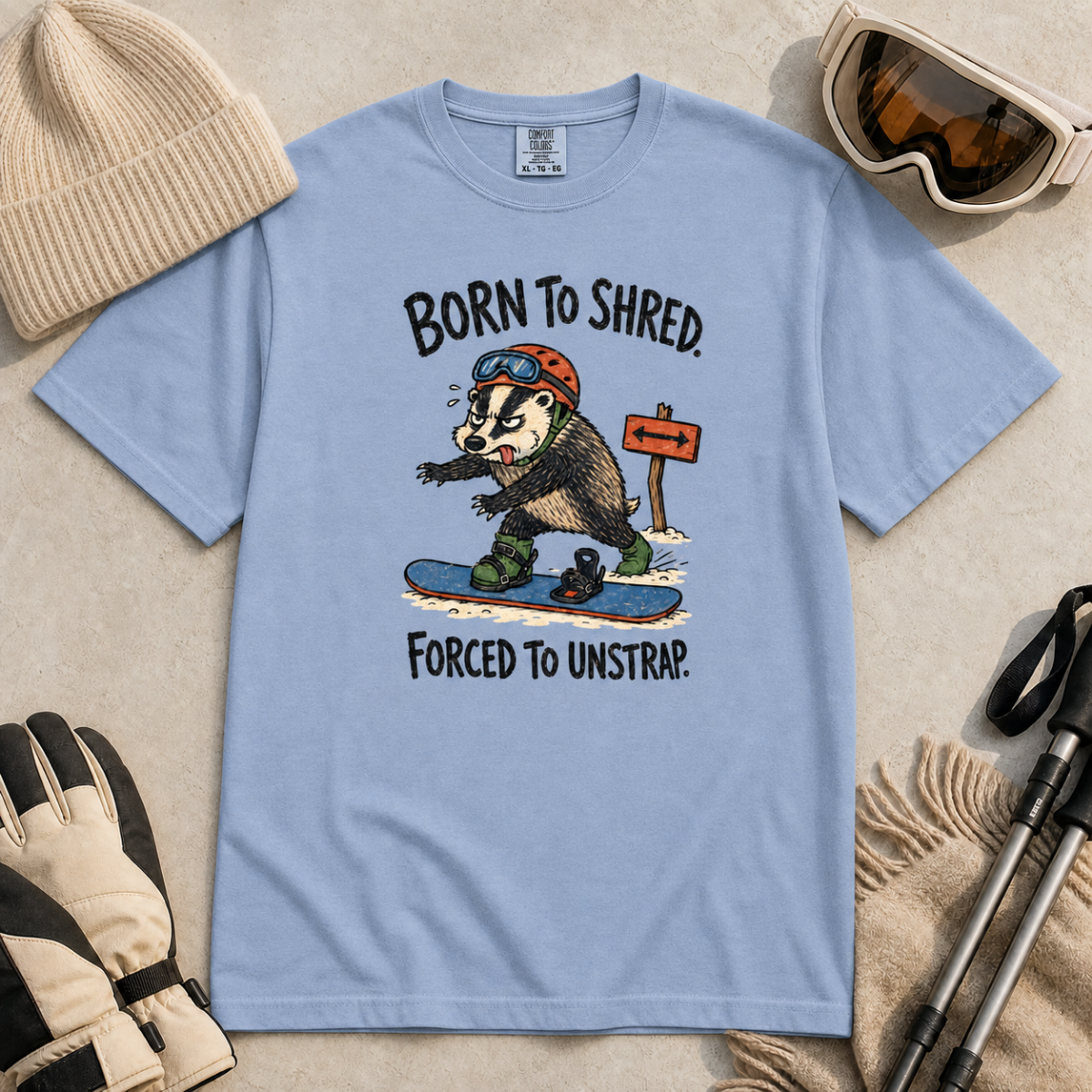 Flat lay of a washed denim blue t-shirt featuring a frustrated badger snowboarder removing one foot from its binding, with “Born to Shred. Forced to Unstrap.” text.