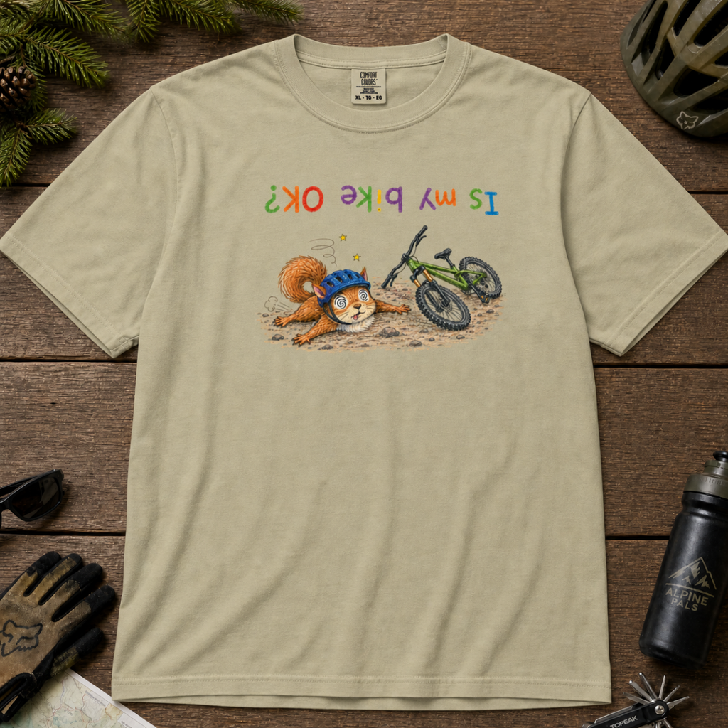 Flat lay of a khaki colored t-shirt featuring a humorous squirrel mountain bike crash design with an upside-down “Is My Bike OK?” message.