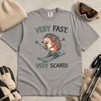 Flat lay of a gray t-shirt featuring a frightened hedgehog skiing at high speed with “Very Fast. Very Scared.” text, styled with winter accessories.