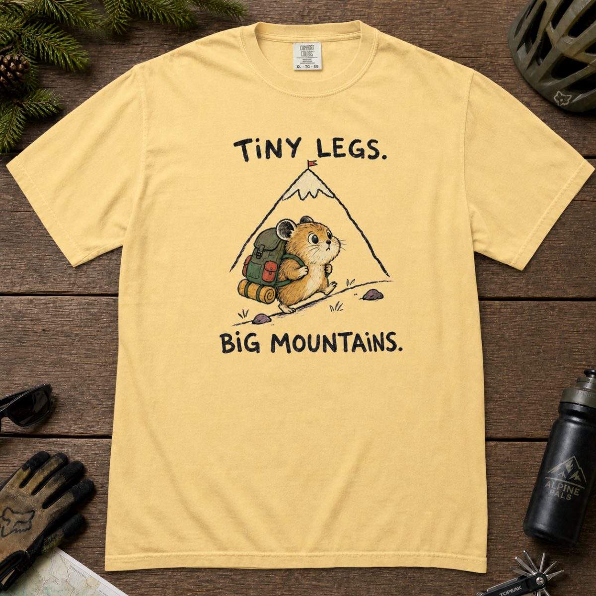 Flat lay of a butter yellow t-shirt with a playful mountain hiking design showing a small backpacking hamster approaching a tall peak.