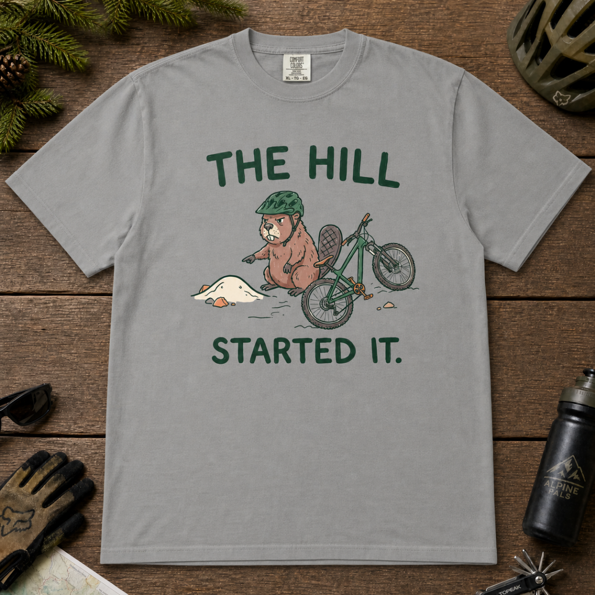 Flat lay of a grey t-shirt with a humorous mountain biking design showing an annoyed beaver blaming a dirt hill beside its bike.