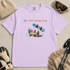 Flat lay of an orchid t-shirt featuring an upside-down “Is My Snowboard OK?” design with a dazed otter lying in the snow beneath a loose snowboard.