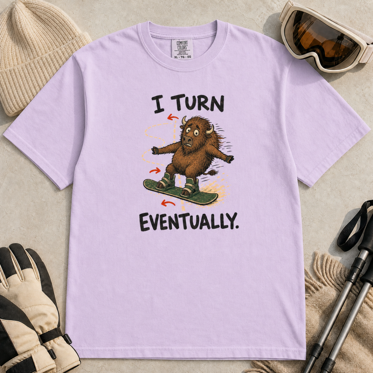Flat lay of an orchid t-shirt featuring a nervous bison snowboarding along a winding path with “I Turn Eventually.” text.