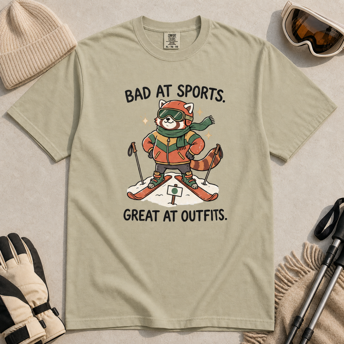 Flat lay of a khaki t-shirt with a humorous red panda ski design celebrating fashionable gear over athletic ability.
