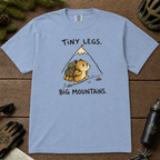 Flat lay of a washed denim blue t-shirt with a playful mountain hiking design showing a small backpacking hamster approaching a tall peak.