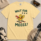 Flat lay of a butter yellow t-shirt featuring a panicked duck skiing behind the group with a red scarf and “Wait for Meeee!” text.