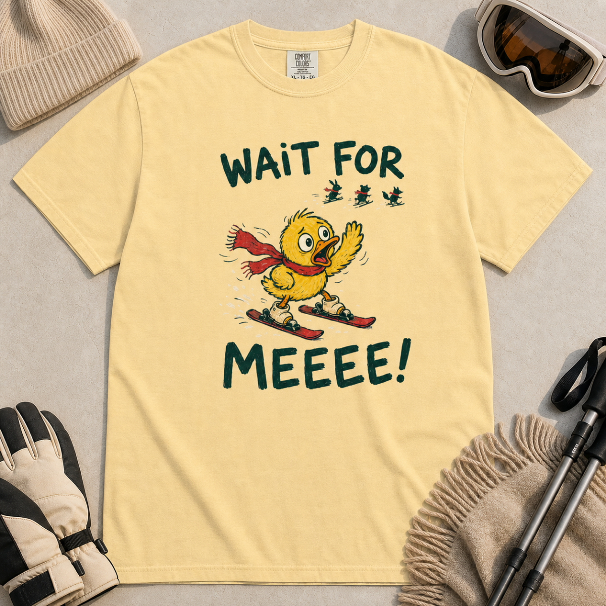 Flat lay of a butter yellow t-shirt featuring a panicked duck skiing behind the group with a red scarf and “Wait for Meeee!” text.