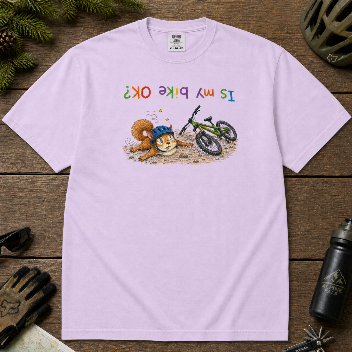 Flat lay of an orchid colored t-shirt featuring a humorous squirrel mountain bike crash design with an upside-down “Is My Bike OK?” message.