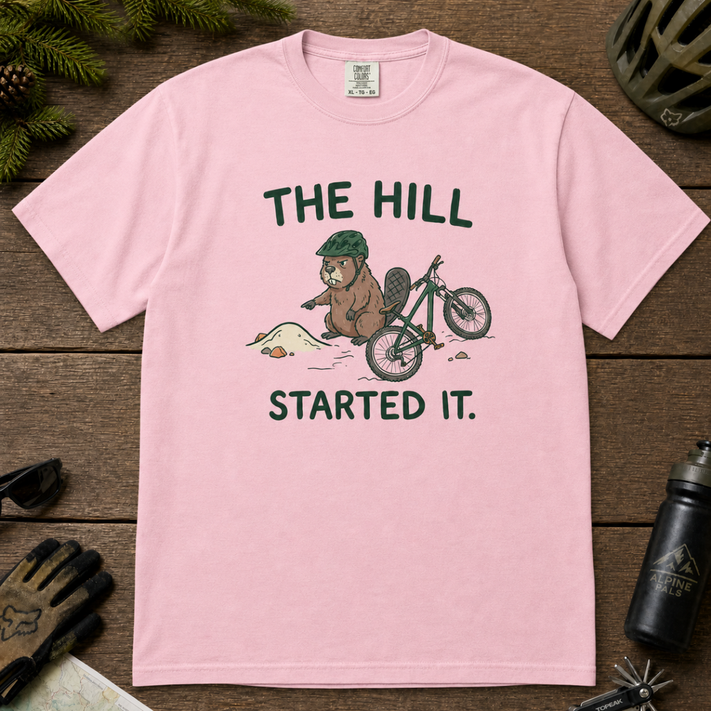 Flat lay of a blossom pink t-shirt with a humorous mountain biking design showing an annoyed beaver blaming a dirt hill beside its bike.