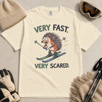 Flat lay of an ivory white t-shirt featuring a frightened hedgehog skiing at high speed with “Very Fast. Very Scared.” text, styled with winter accessories.
