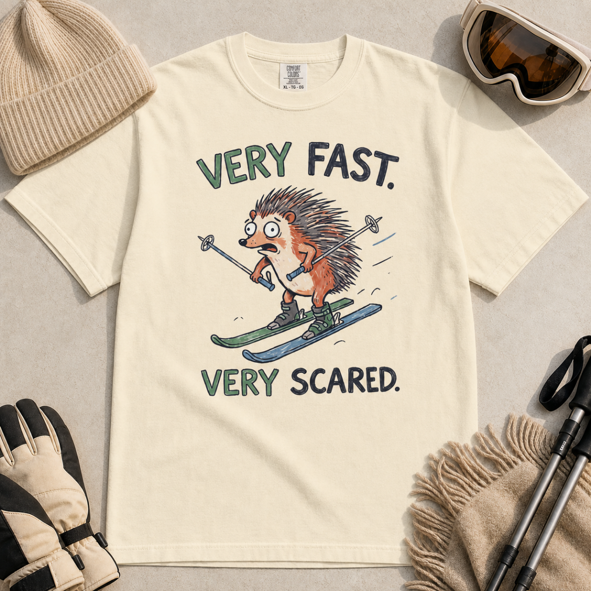 Flat lay of an ivory white t-shirt featuring a frightened hedgehog skiing at high speed with “Very Fast. Very Scared.” text, styled with winter accessories.
