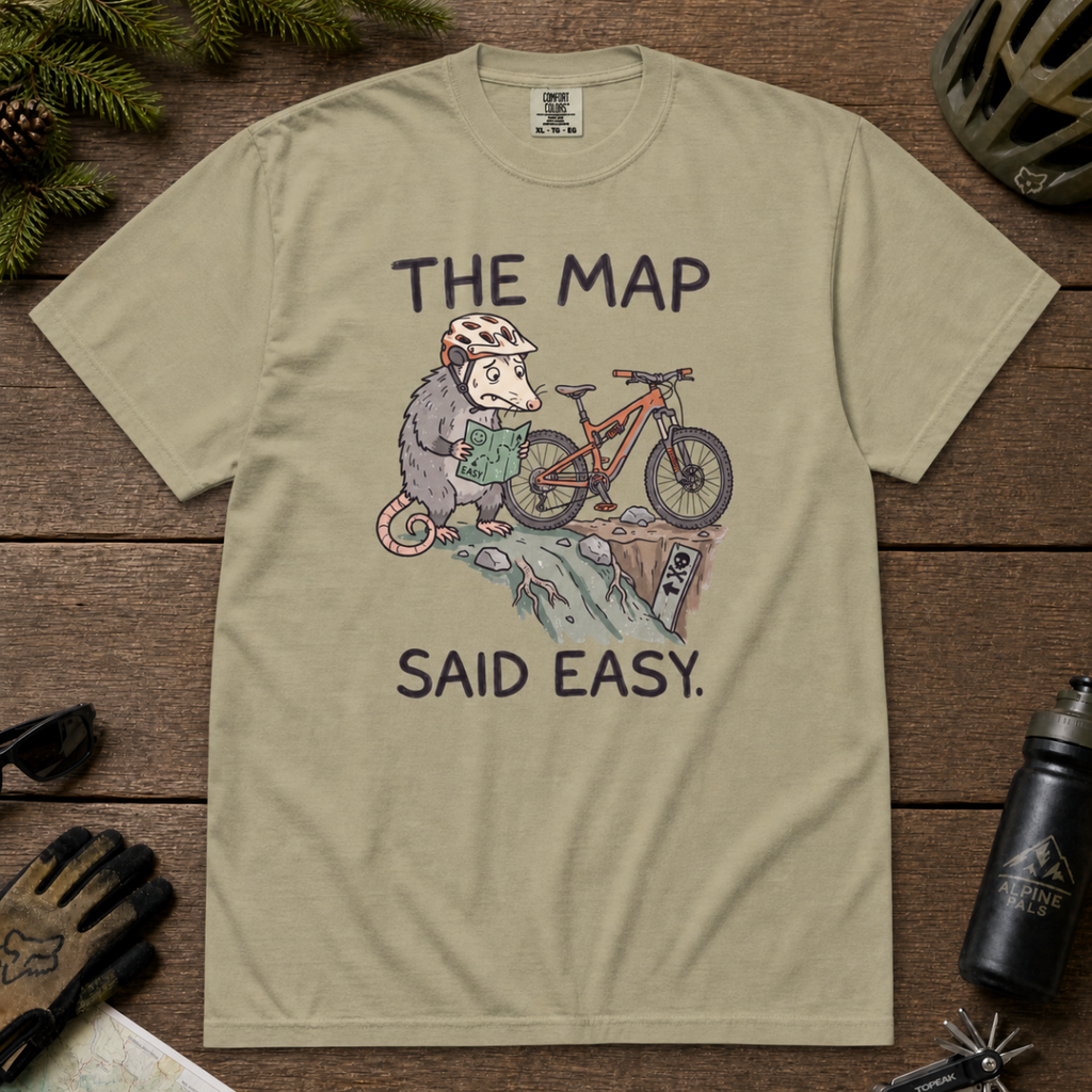 Flat lay of a khaki t-shirt with a humorous mountain biking design showing a nervous opossum, an orange bike, and a dangerously steep trail.