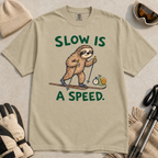 Flat lay of a khaki t-shirt with a humorous sloth skiing graphic, a stopwatch-holding snail, and bold green “Slow Is a Speed.” text.