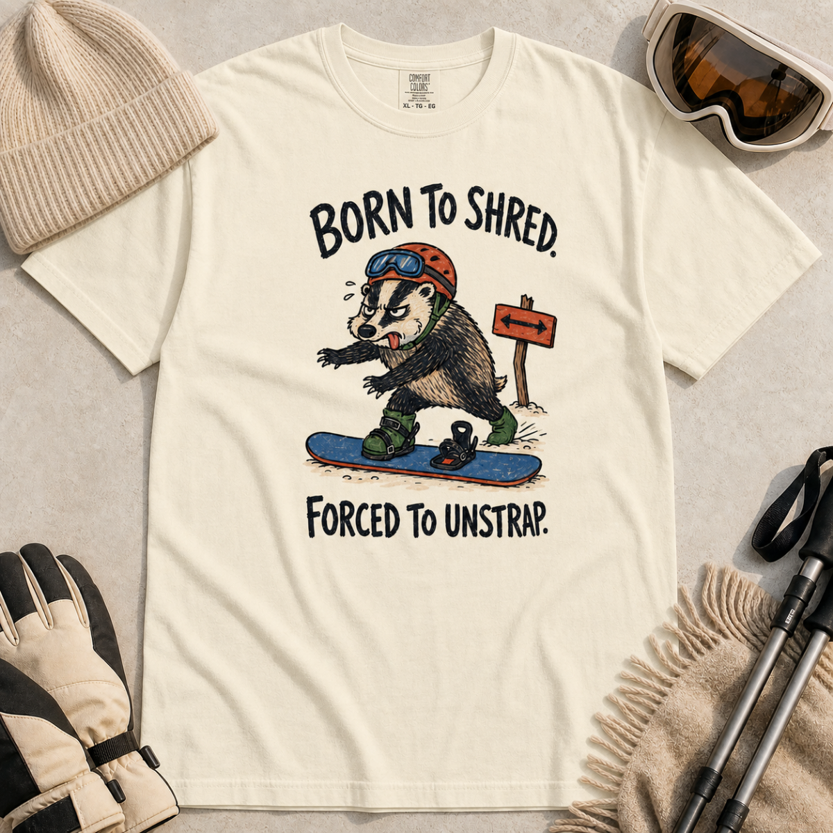 Flat lay of an ivory white t-shirt featuring a frustrated badger snowboarder removing one foot from its binding, with “Born to Shred. Forced to Unstrap.” text.