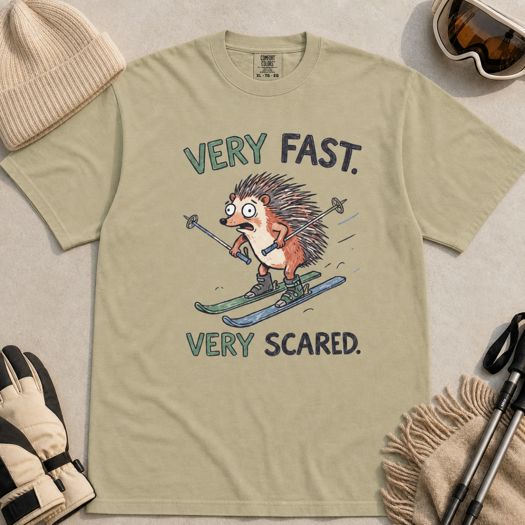 Flat lay of a khaki t-shirt featuring a frightened hedgehog skiing at high speed with “Very Fast. Very Scared.” text, styled with winter accessories.