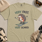 Flat lay of a khaki t-shirt featuring a frightened hedgehog skiing at high speed with “Very Fast. Very Scared.” text, styled with winter accessories.