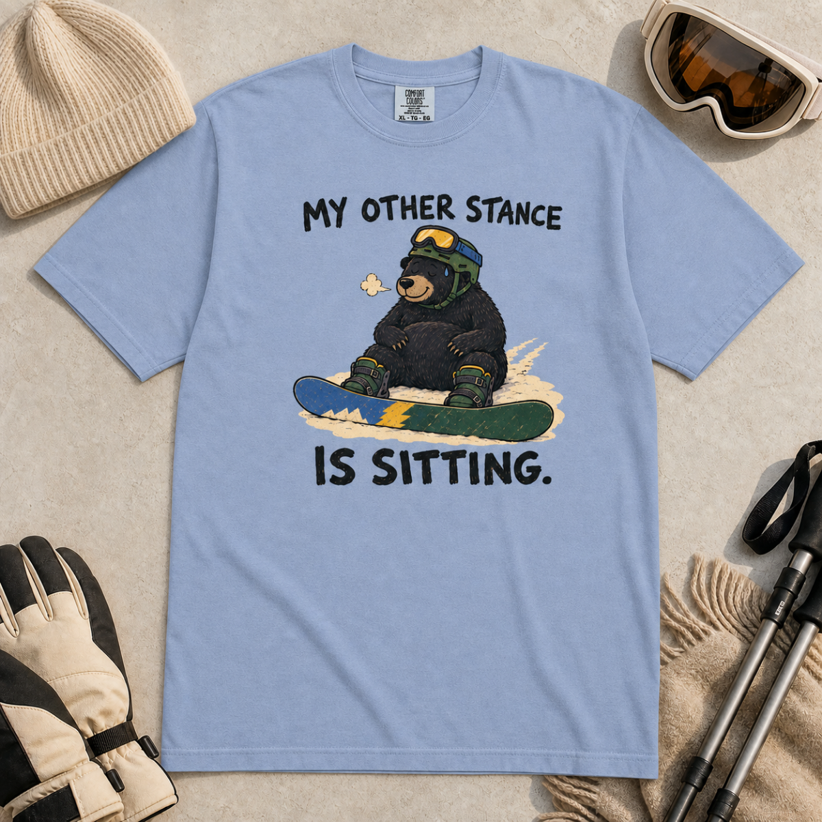 Flat lay of a washed denim blue t-shirt with a humorous snowboarding design showing a resting bear seated behind its board.