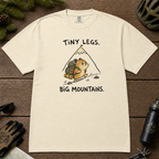 Flat lay of an ivory white t-shirt with a playful mountain hiking design showing a small backpacking hamster approaching a tall peak.